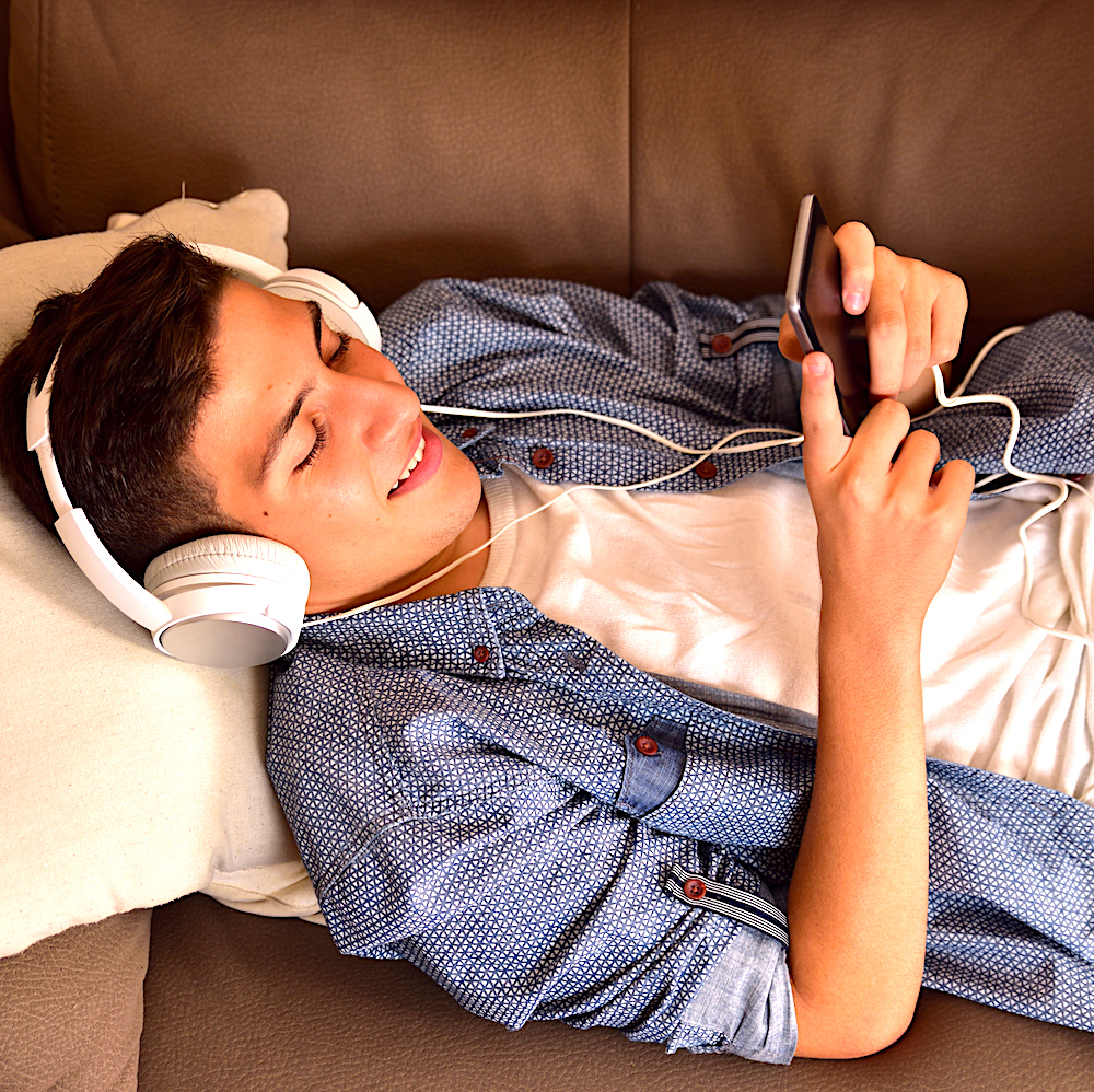 Teen lying face up on couch with hardphones and smartphone consuming multimedia content top view
