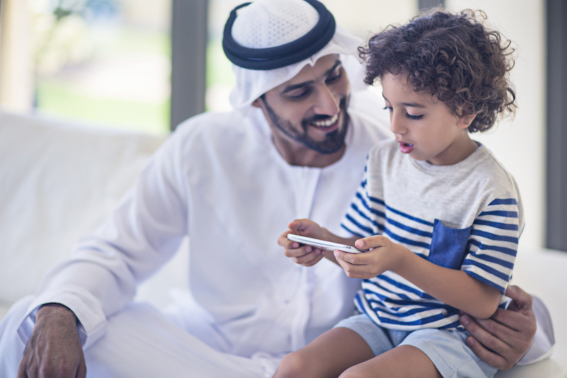Middle Eastern father sharing time with his son at home, kid is playing with the mobile phone while his father is looking at him,the father is wearing the traditional Dishdasha.